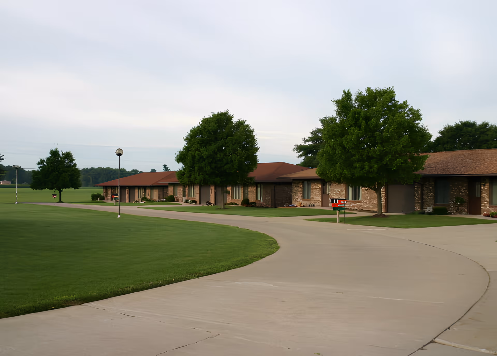A curved concrete driveway leads past a row of single-story brick buildings with brown roofs, surrounded by well-maintained green lawns and several large trees under a cloudy sky.