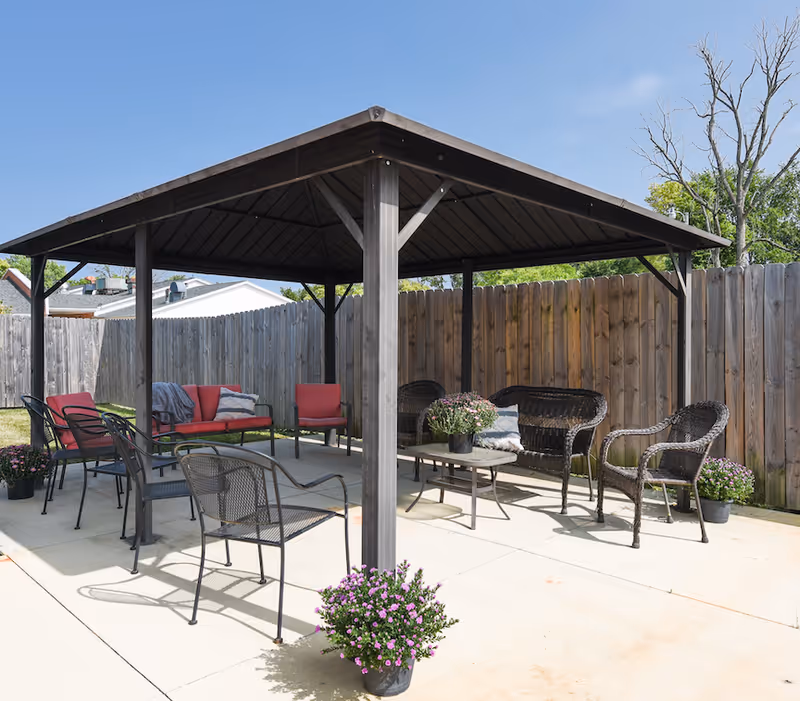 Covered outdoor patio with metal gazebo, seating including chairs and wicker sofas, a coffee table, and potted flowers beside a wooden fence.