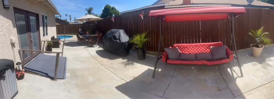 Sunlit fenced patio with a red cushioned swing, covered grill, patio dining set and a small pool next to the building.