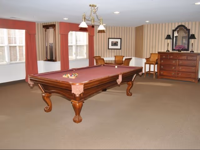 A well-lit room with a pool table in the center featuring a burgundy felt surface and wooden legs. The room has beige carpet and striped wallpaper with a chair rail. There are two windows with red curtains, a wooden dresser with a mirror and two lamps on top, and a small table with two chairs in the corner.