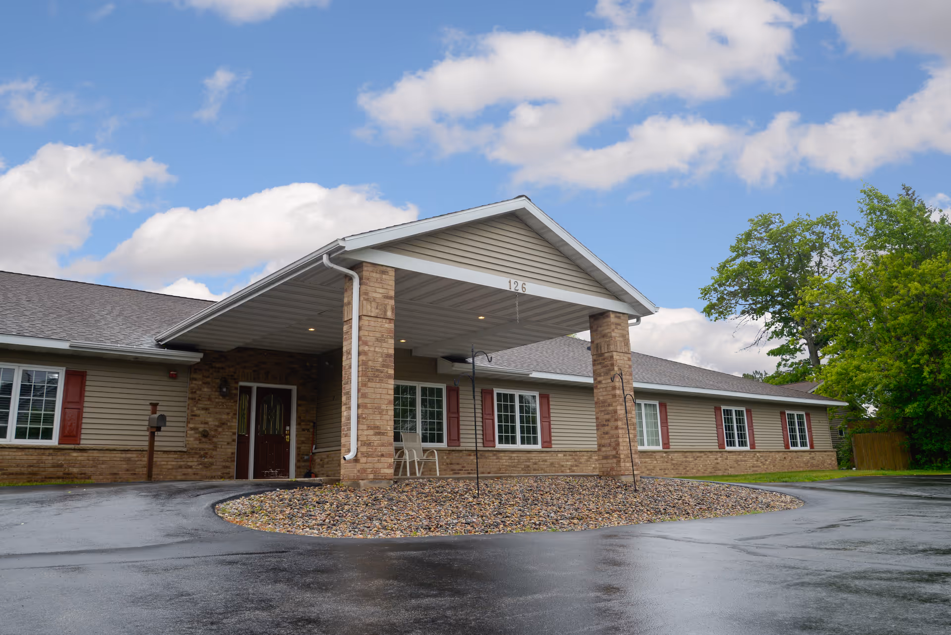 Front entrance of a single-story building with a covered porte-cochère, brick columns, and a circular driveway under a partly cloudy sky.