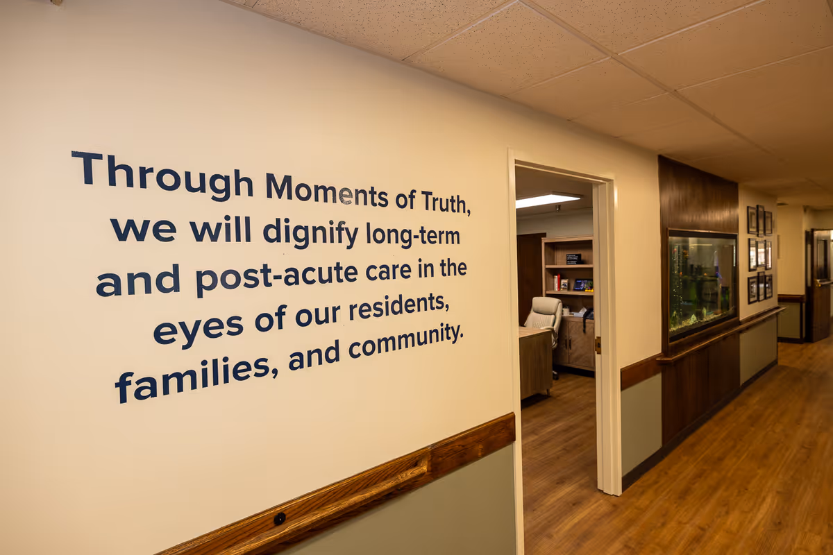 Interior hallway of a senior living facility with a motivational quote on the wall that reads: 'Through Moments of Truth, we will dignify long-term and post-acute care in the eyes of our residents, families, and community.' The hallway has wood flooring, a wooden handrail, and an aquarium built into the wall. An open door reveals an office with a chair and shelves.