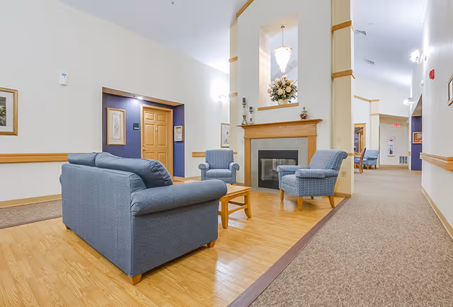 A bright communal lounge with blue upholstered sofas and chairs arranged around a fireplace in a hallway of a senior living facility.
