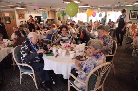 A large group of elderly people seated at round tables in a decorated dining room, enjoying a meal together. The room is adorned with colorful balloons and festive decorations. Several people are wearing leis, and a server is walking through the room. The atmosphere appears lively and social.