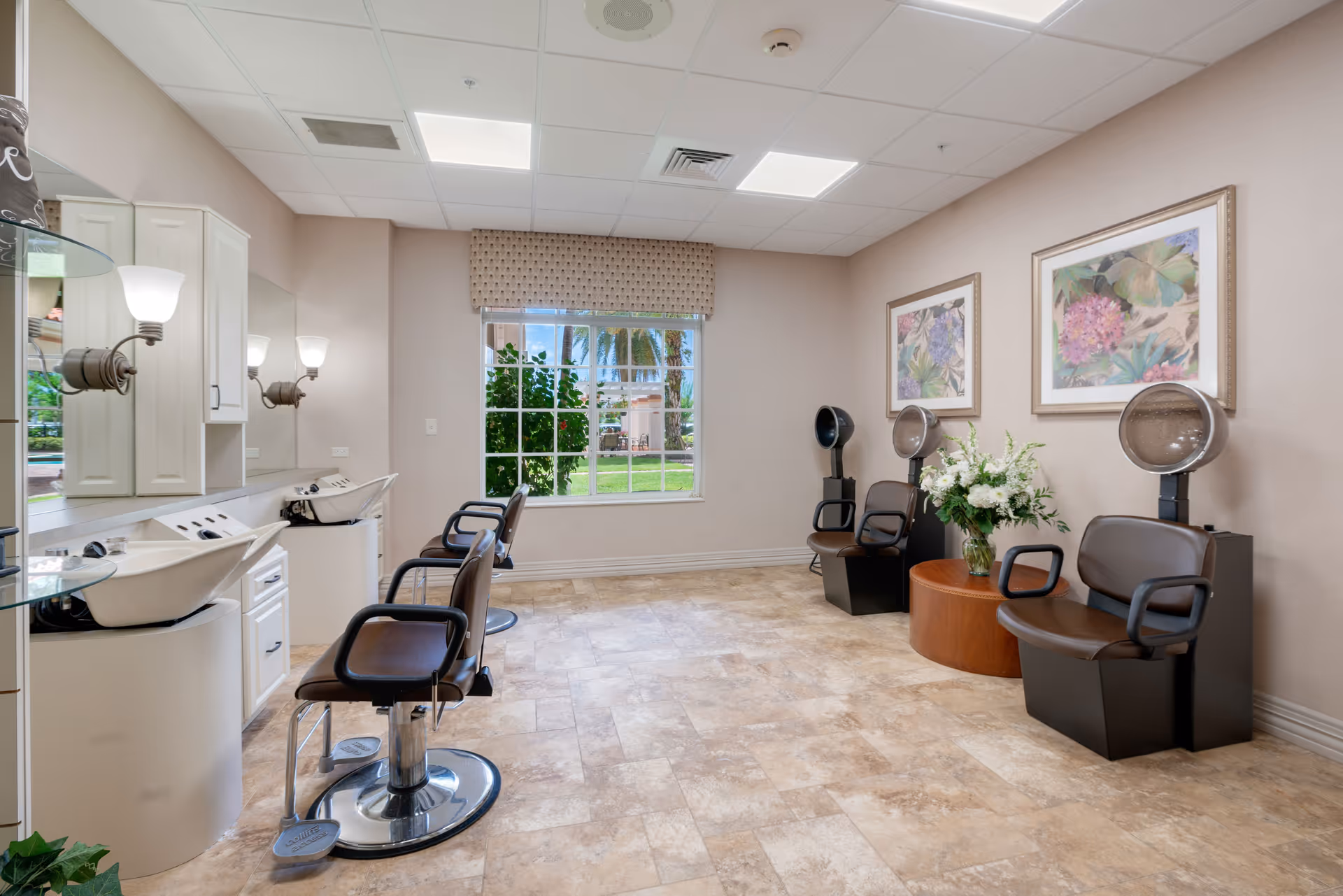 A bright and clean hair salon area inside a senior living facility. The room features three salon chairs with sinks for washing hair on the left side, and three hair drying chairs with hooded dryers on the right side. There is a large window in the center of the back wall letting in natural light, with a view of greenery and palm trees outside. The walls are decorated with framed floral artwork, and a round wooden table with a vase of white flowers is placed between the drying chairs. The floor is tiled, and the ceiling has recessed lighting panels.