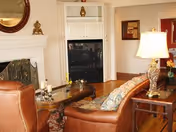 A cozy living room with a brown leather sofa and armchair, a wooden coffee table with candles and decorative items, a fireplace with a round mirror above it, a built-in white shelving unit with a TV, and a table lamp on a side table next to the sofa.
