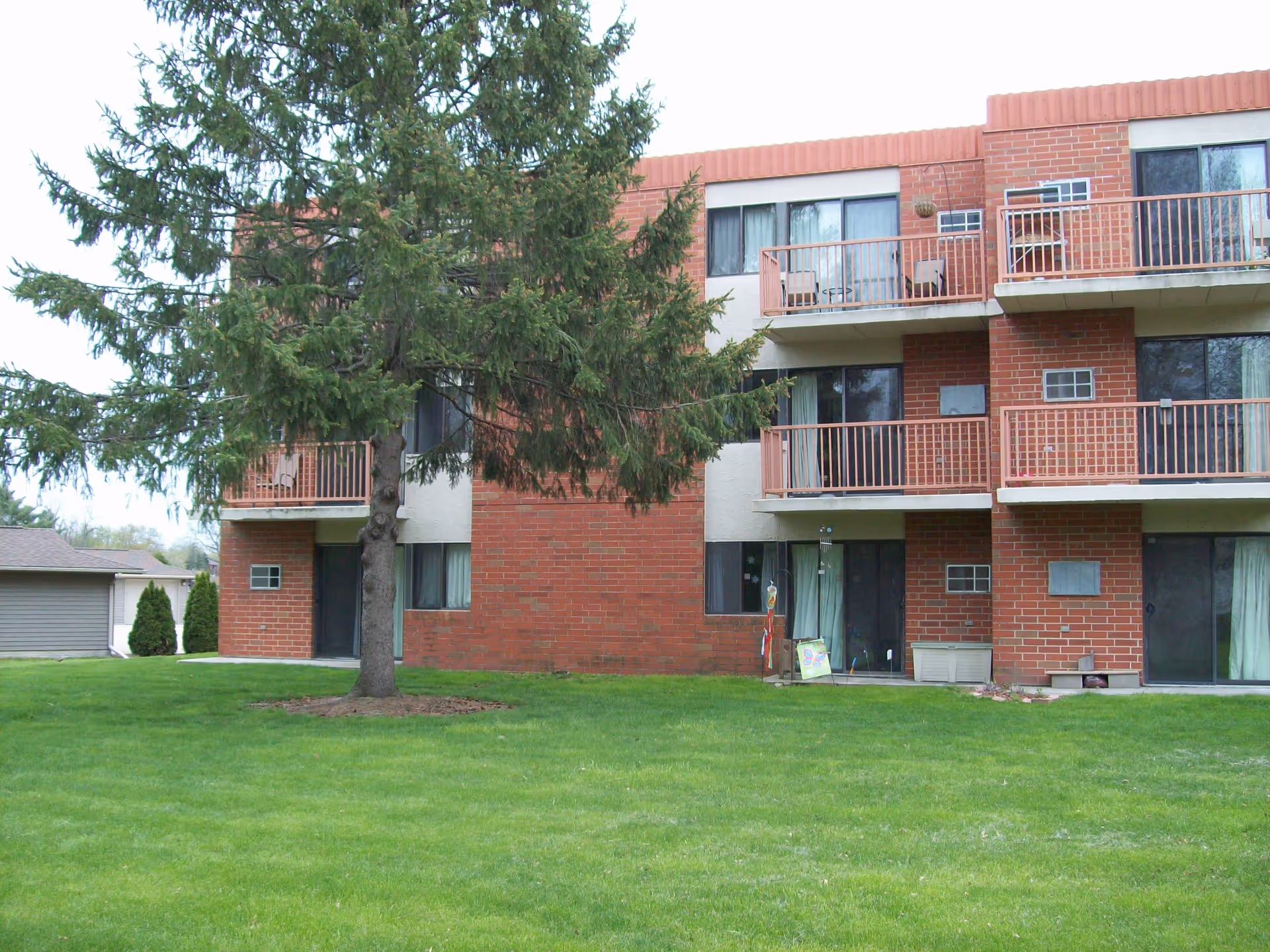 Exterior view of a three-story brick senior living building with balconies and sliding glass doors. A large evergreen tree stands in front of the building on a well-maintained green lawn.