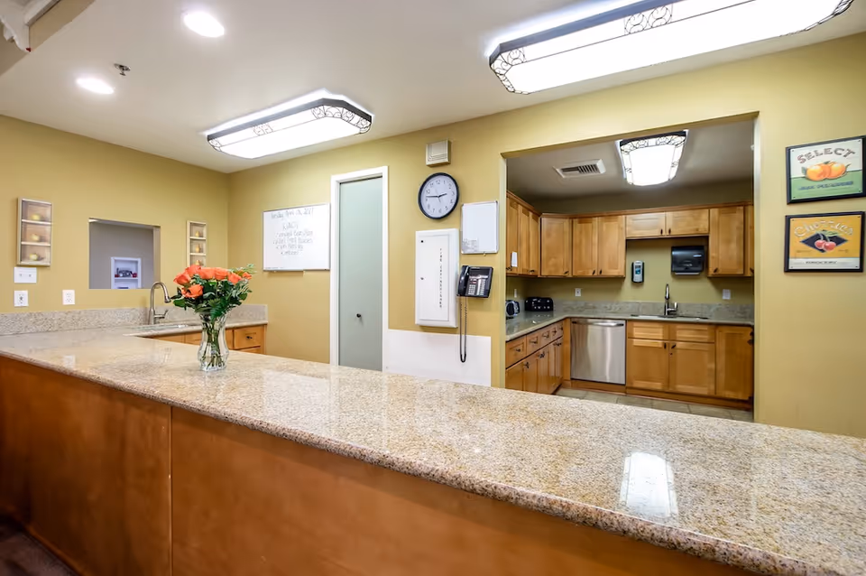 Bright communal kitchen and service counter with a long granite countertop, a vase of flowers, wooden cabinets and a stainless dishwasher.