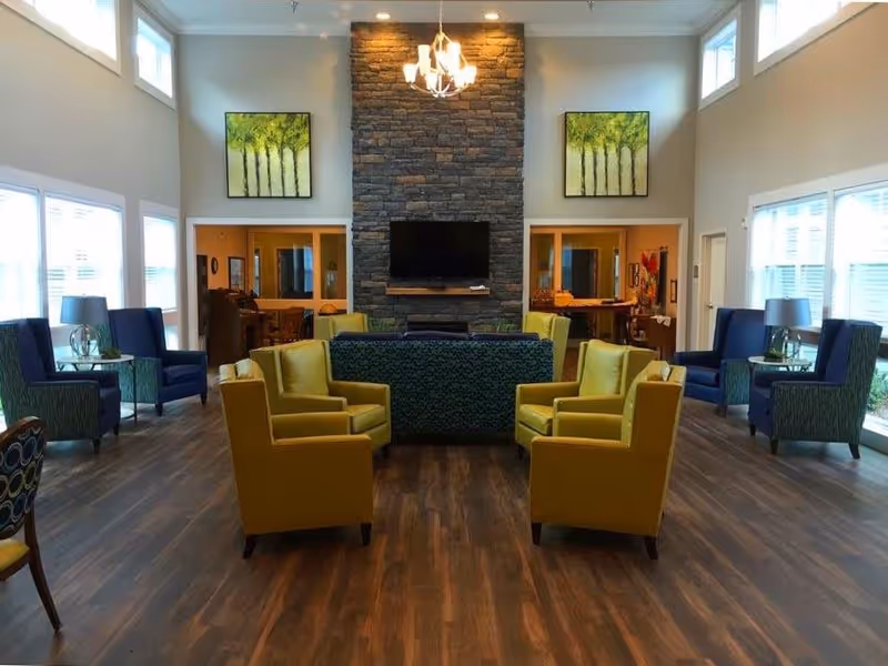 A spacious living room with wooden flooring, featuring a central stone fireplace with a mounted flat-screen TV. The room is furnished with yellow armchairs, blue armchairs, and a patterned sofa. Two large paintings of trees hang on the wall above the fireplace. Large windows on both sides allow natural light to fill the room.