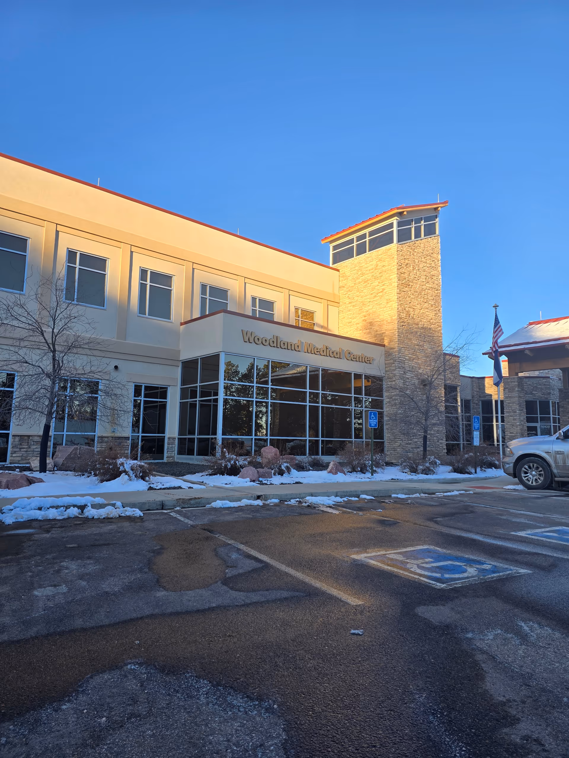 Exterior view of Woodland Medical Center building with large windows and stone tower under a clear blue sky. There is snow on the ground and several handicap parking spaces in front of the building.