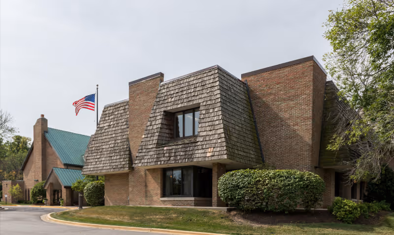 Exterior view of a senior living facility building with brick walls and a shingled roof section. There is an American flag on a flagpole near the building, surrounded by trimmed bushes and trees under a clear sky.