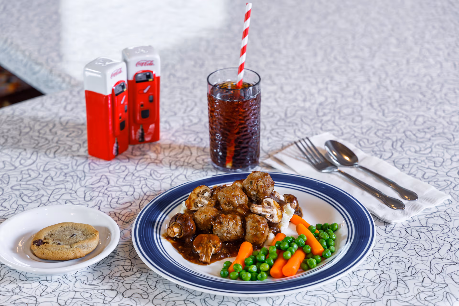 Plate of meatballs with mushrooms, peas and carrots on a diner-style table with a cookie, a glass of cola with a striped straw, utensils, and vintage Coca-Cola salt-and-pepper shakers.