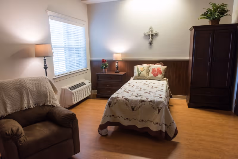 Sunlit furnished bedroom with a single bed, nightstand and lamp, armchair, wardrobe, and a decorative cross on the wall.