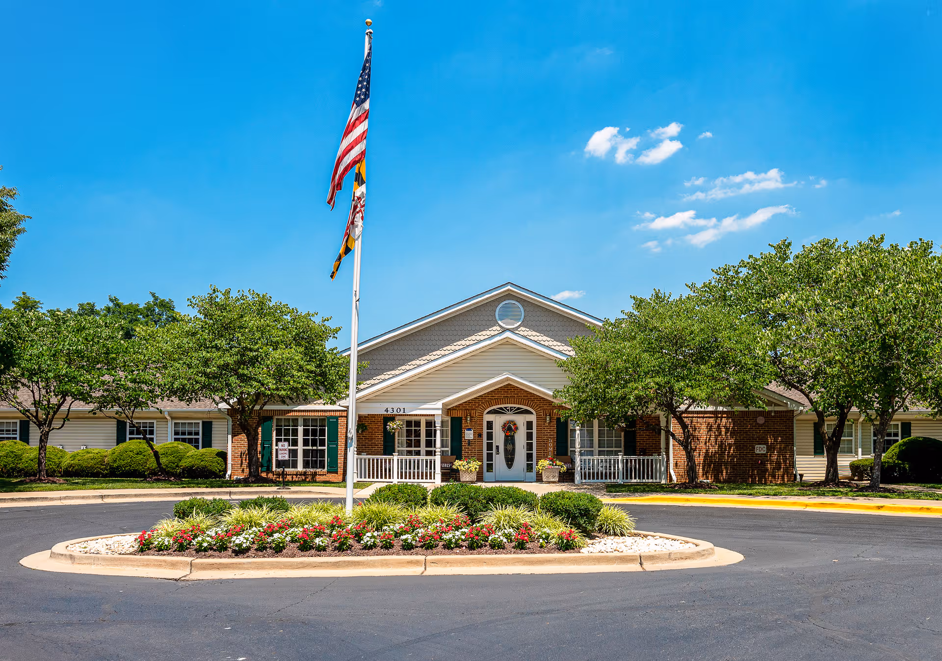 Front entrance of a single-story brick senior living community with a flagpole and a landscaped circular driveway.