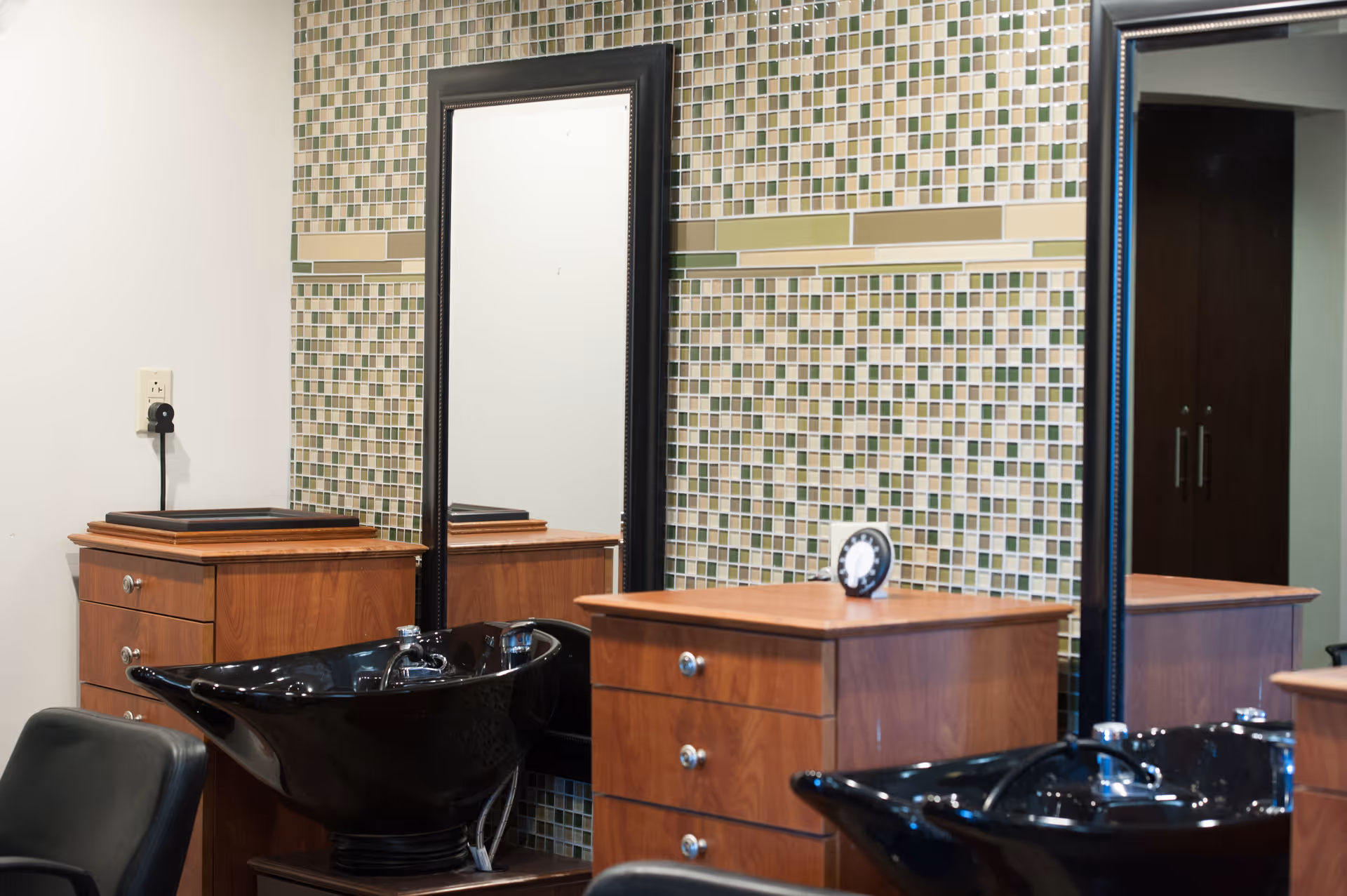 Salon-style hair-washing stations with black sinks, wooden cabinetry and large mirrors against a mosaic-tiled wall.