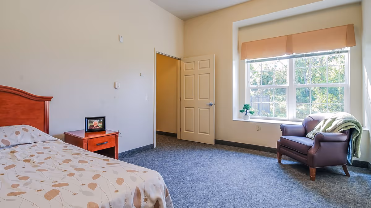 A bright senior living bedroom with a single bed covered in a patterned beige and brown bedspread, a wooden nightstand with a framed photo, an open door leading to another room, a large window with a beige valance, a small potted plant on the windowsill, and a brown armchair with a green blanket draped over one arm.