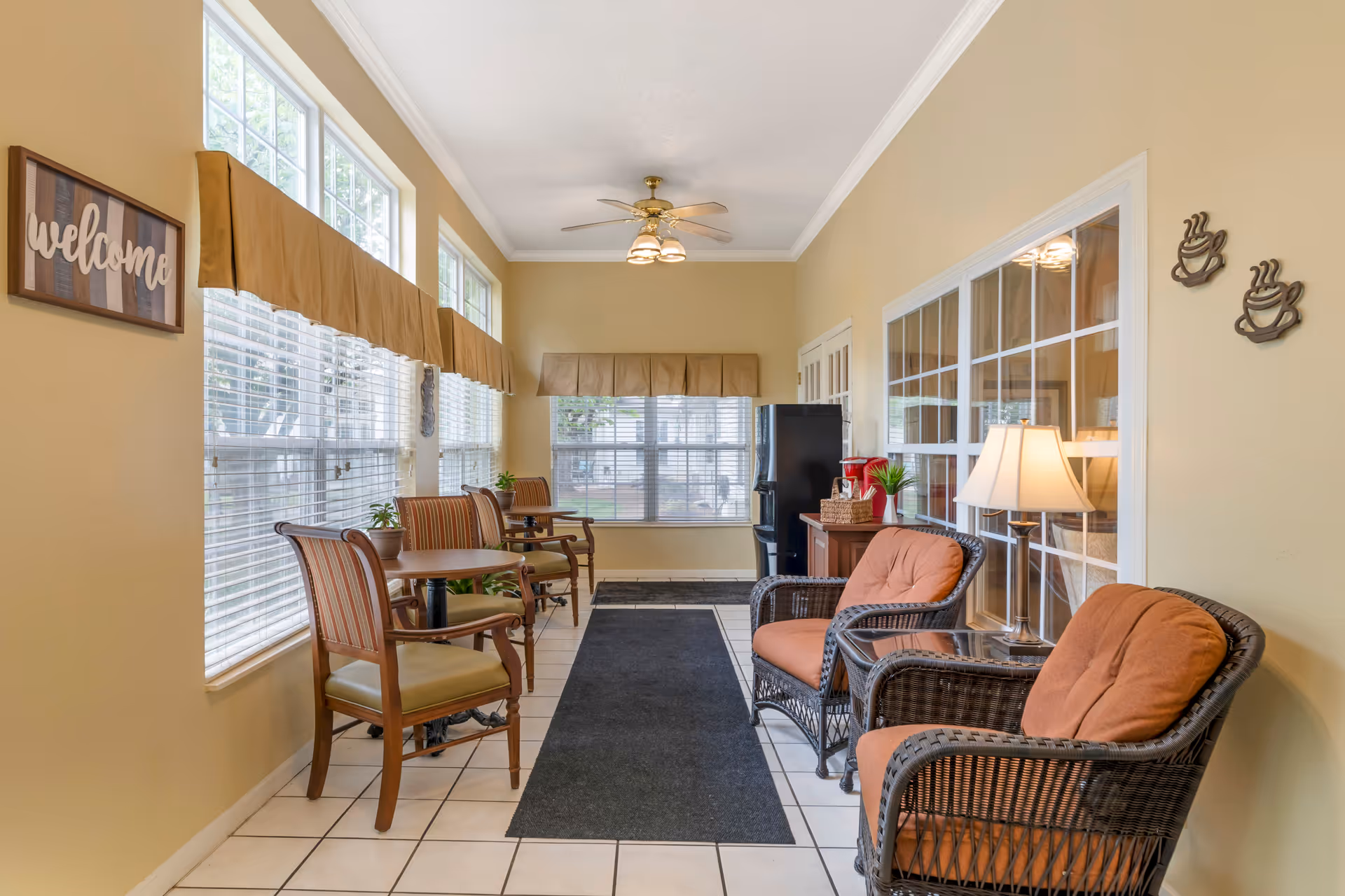 A bright, cozy sitting area with large windows covered by beige valances. The room features two wicker armchairs with orange cushions, a small table with a lamp between them, and two wooden chairs with striped cushions paired with small round tables. A black refrigerator and a red coffee maker are visible in the background. The walls are painted light yellow, and there is a 'welcome' sign on the left wall and two coffee cup wall decorations on the right.
