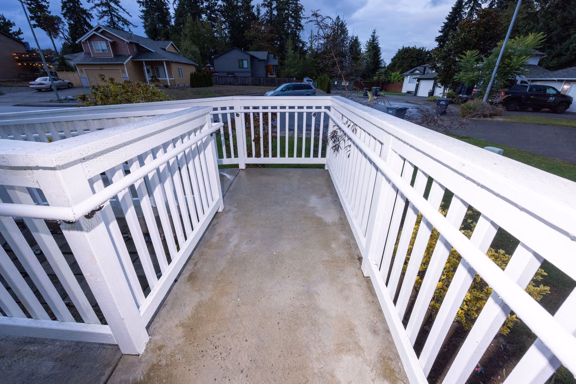 A white wooden ramp with handrails on both sides leading down to a residential street with houses and parked cars, surrounded by trees and greenery under a cloudy sky.