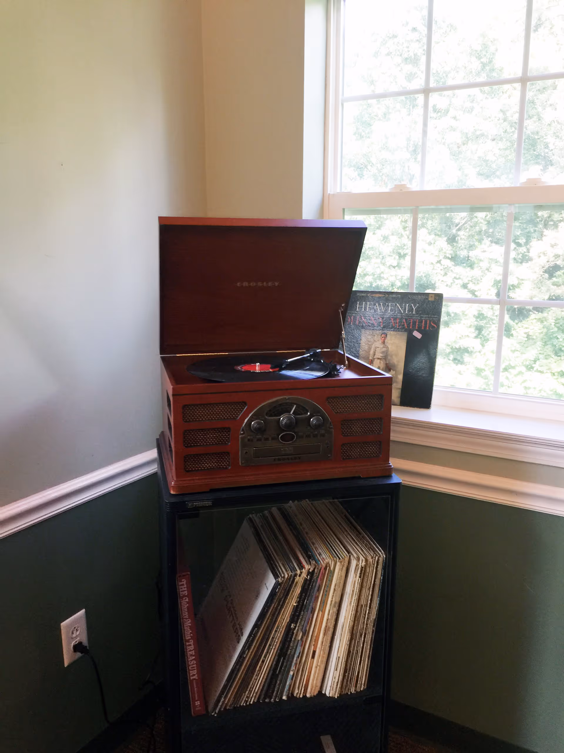 A vintage Crosley record player with a vinyl on the turntable sits on a cabinet above a shelf of records beside a large window.