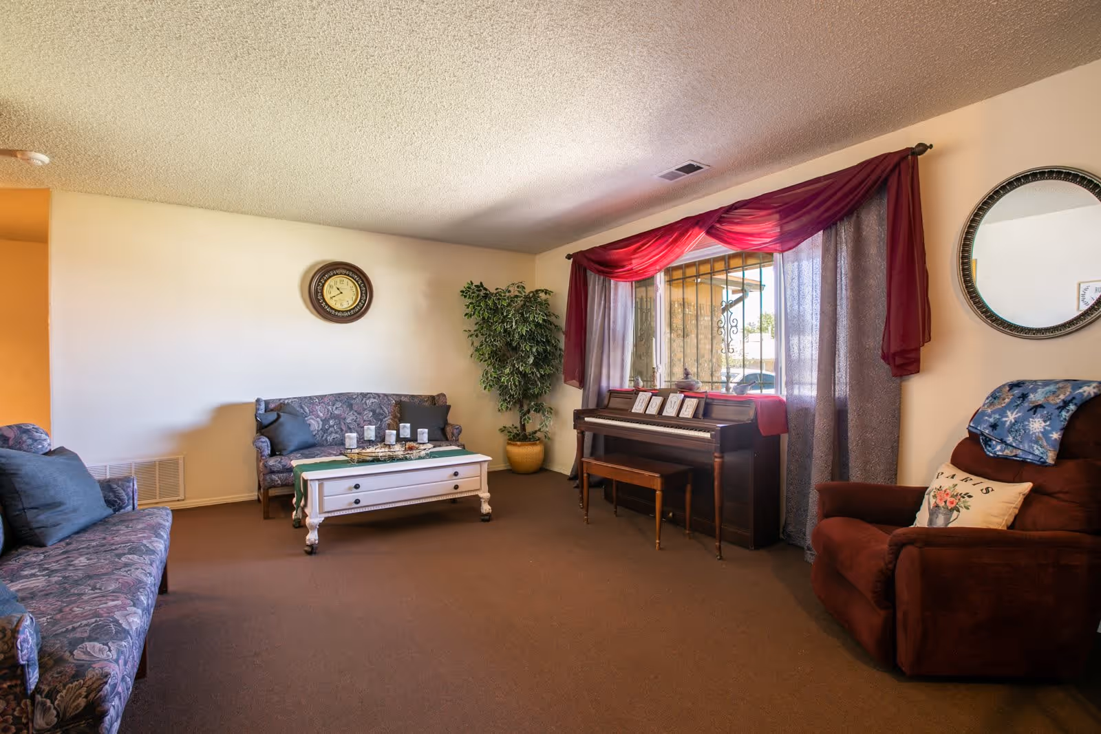 A cozy living room with two floral patterned sofas, a white coffee table with candles, a brown recliner chair with a floral pillow and a blue blanket, a piano with framed photos on top, a large window with sheer and red curtains, a round wall clock, a round mirror, and a potted plant in the corner.