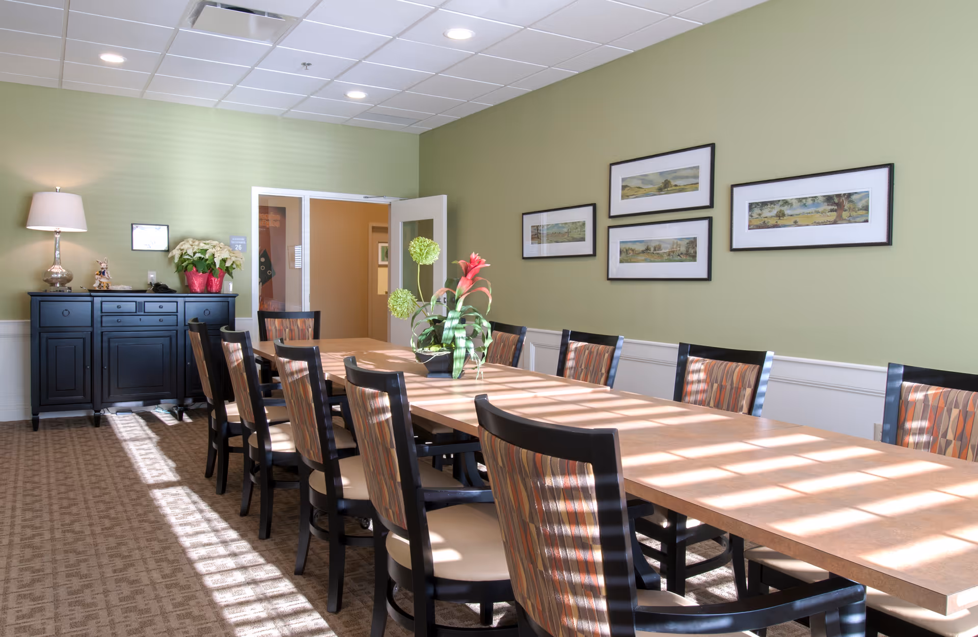 A well-lit dining room with a long rectangular table surrounded by chairs with patterned upholstery. The walls are painted light green and decorated with four framed landscape paintings. A black sideboard with a lamp, a potted plant, and decorative items is against one wall. Sunlight streams through windows, casting shadows on the table and carpeted floor.