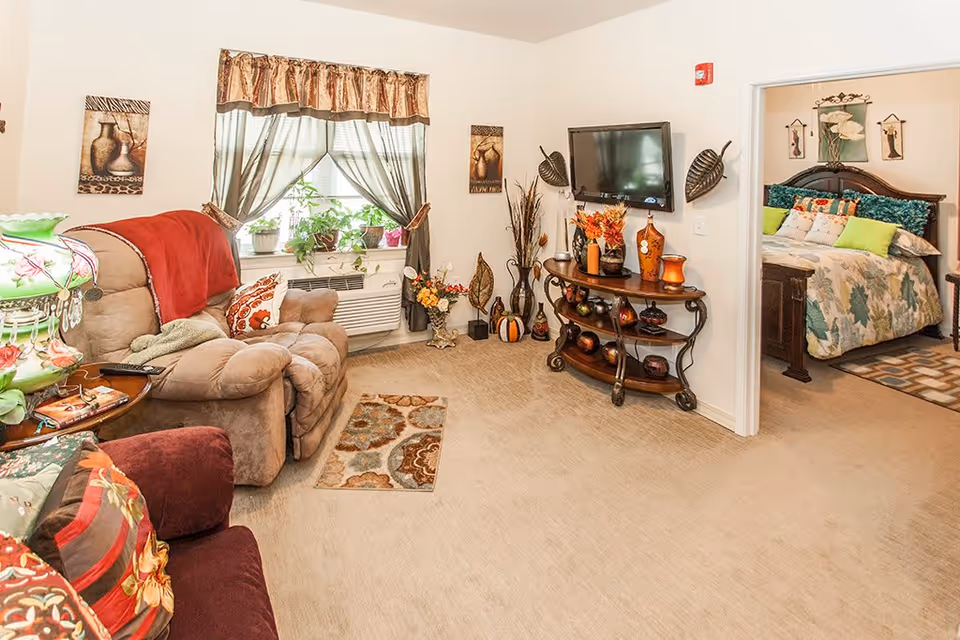 A cozy living room with beige carpet, a recliner chair with a red throw blanket, a maroon armchair with colorful cushions, a window with sheer curtains and potted plants on the windowsill, a wall-mounted flat-screen TV above a decorative wooden console table with vases and autumn-themed decor. An adjacent bedroom is visible with a bed covered in a floral quilt and multiple pillows, along with wall art above the headboard.