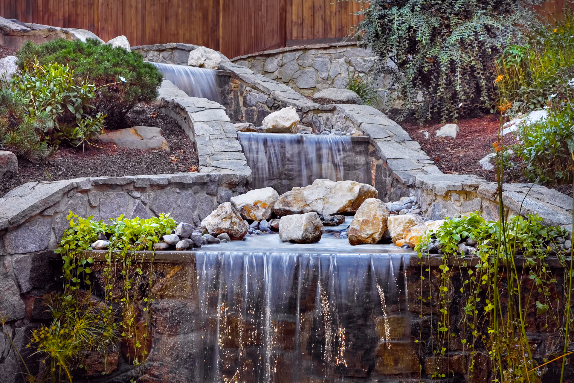 A serene outdoor water feature with cascading waterfalls flowing over stone walls surrounded by rocks, green plants, and a wooden fence in the background.