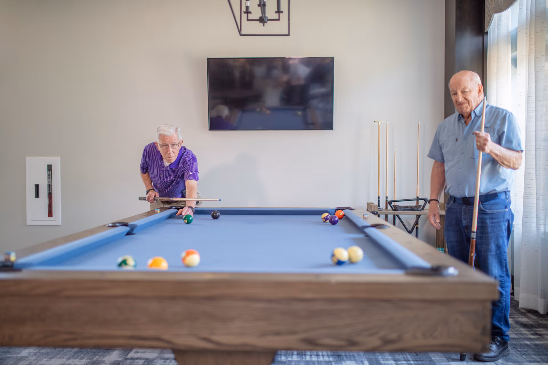 Two elderly men playing pool in a well-lit room with a blue pool table. One man is aiming to take a shot while the other stands nearby holding a cue stick. A flat-screen TV is mounted on the wall behind them, and a rack with additional pool cues is visible near a window with curtains.