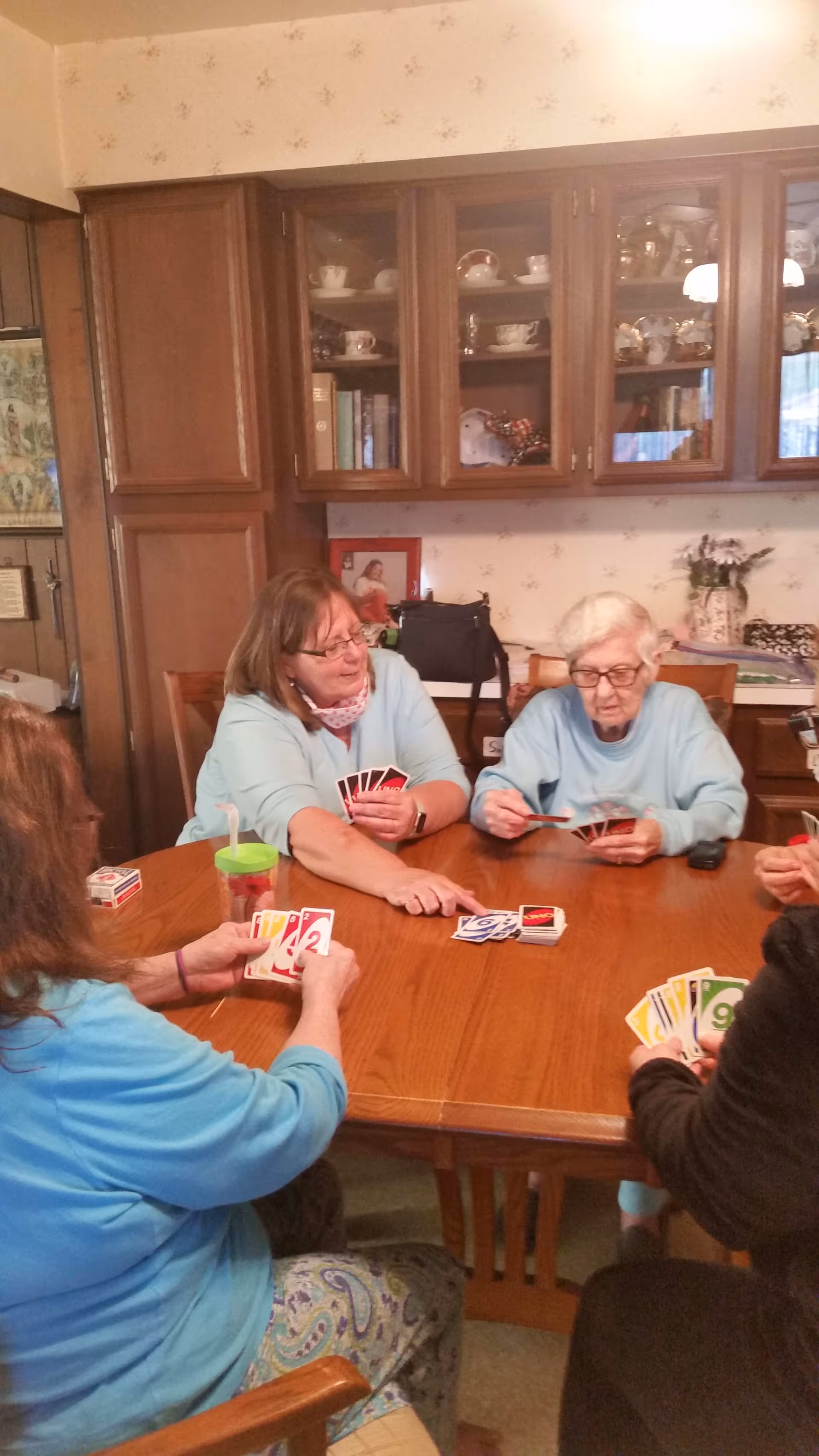 Four elderly women sitting around a wooden table playing a card game in a cozy room with wooden cabinets and shelves displaying dishes and books in the background.