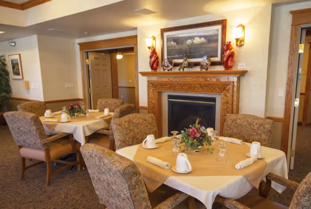 A cozy dining room with round tables covered in beige tablecloths, each set with white mugs, rolled napkins, and glassware. The room features upholstered chairs with floral patterns, a wooden fireplace with decorative vases and a framed painting above it, and warm lighting from wall sconces.
