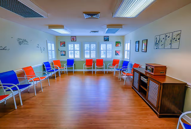 A music activity room featuring colorful chairs arranged in a circle, with large windows allowing natural light, and framed artwork on the walls.