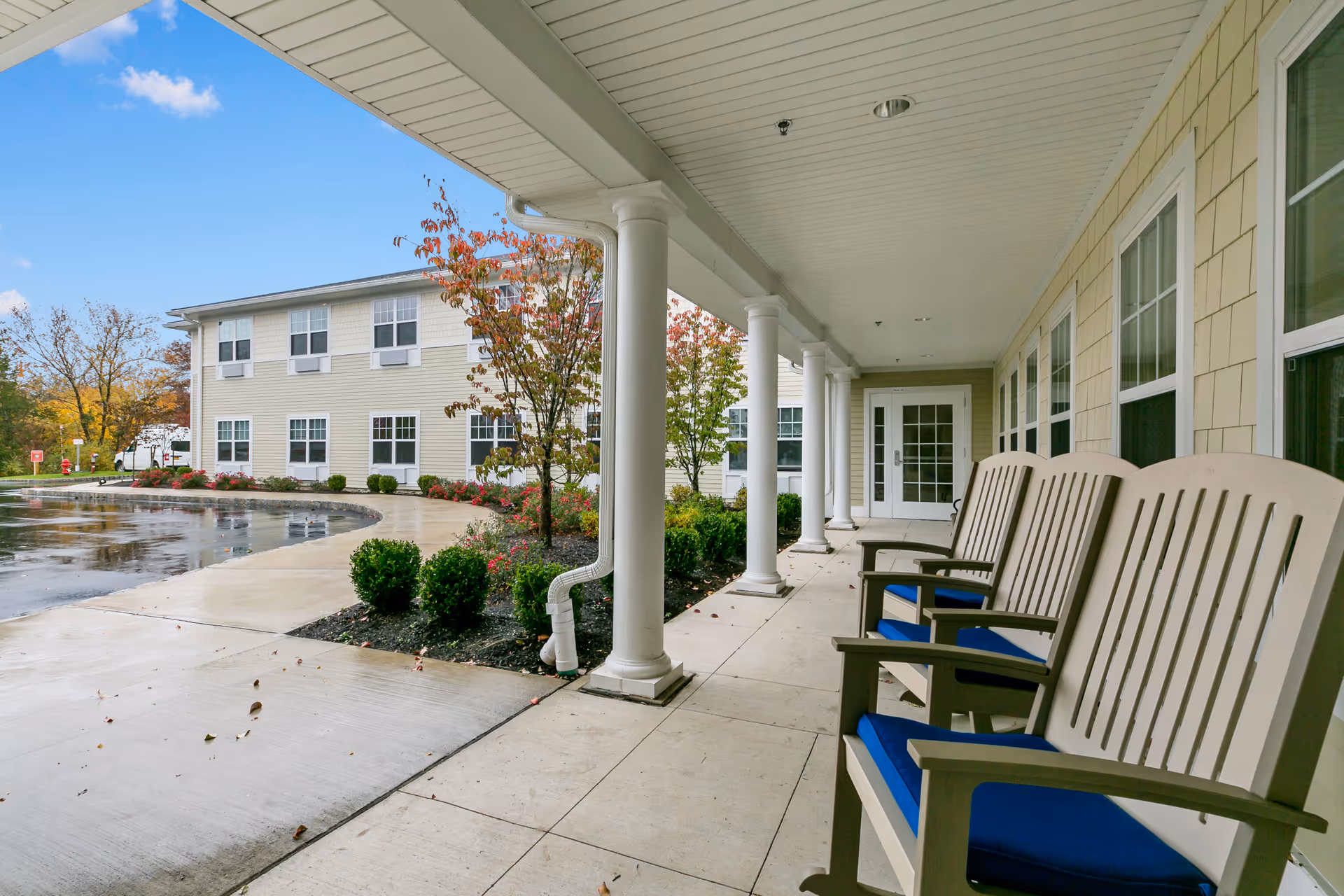 Covered entrance porch with white columns and several outdoor chairs with blue cushions facing the driveway in front of a two-story assisted living building.