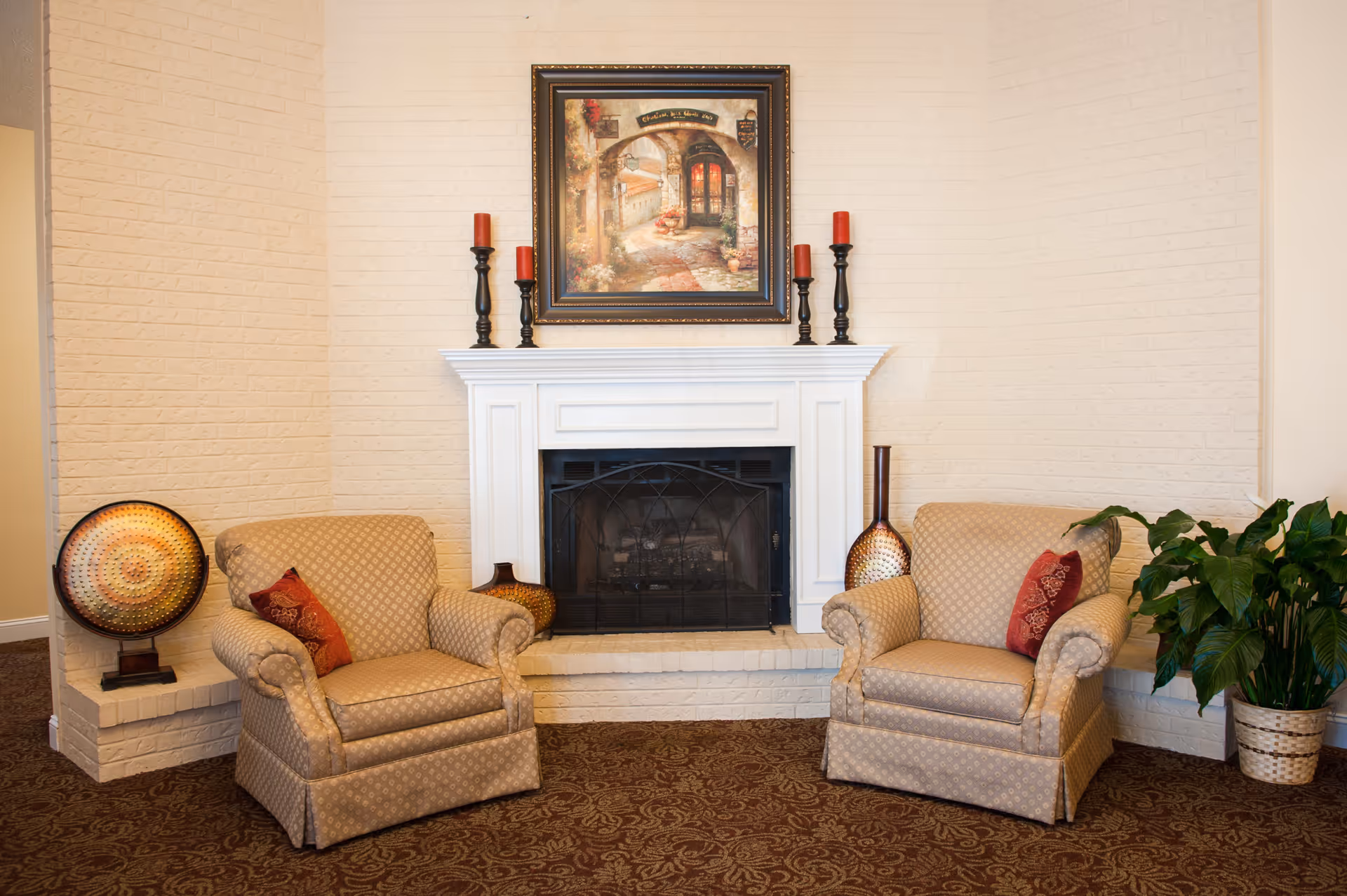 A cozy sitting area with two beige patterned armchairs, each with a red decorative pillow, positioned on either side of a white brick fireplace. The fireplace mantel holds five black candle holders with red candles. Above the mantel is a framed painting depicting a charming outdoor scene. Decorative vases are placed on the hearth, and a green potted plant is on the right side. The walls are light-colored with a textured brick pattern, and the floor is covered with a patterned carpet.