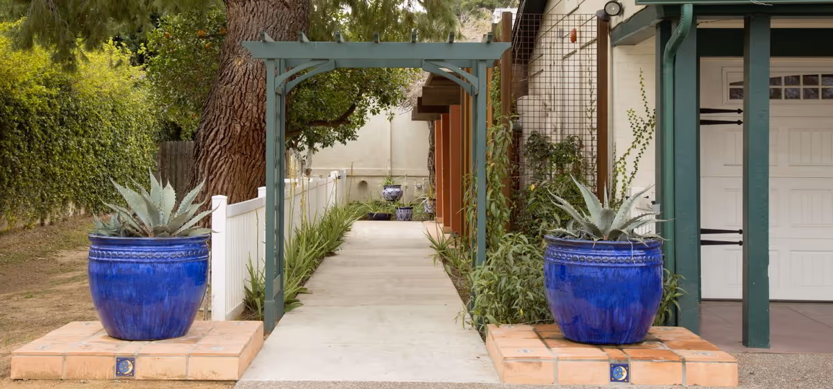 A walkway leading through a garden area with two large blue ceramic pots containing succulent plants on either side. The path is flanked by greenery and a white fence on the left, and a building with green pillars and a garage door on the right. There is a wooden pergola structure over the walkway.