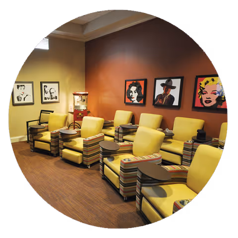 A cozy lounge area with several yellow armchairs featuring striped armrests and attached small round tables. The room has a warm brown carpet and walls painted in beige and rust colors. The wall is decorated with framed pop art portraits of famous personalities. A popcorn machine is placed in the corner near the window.