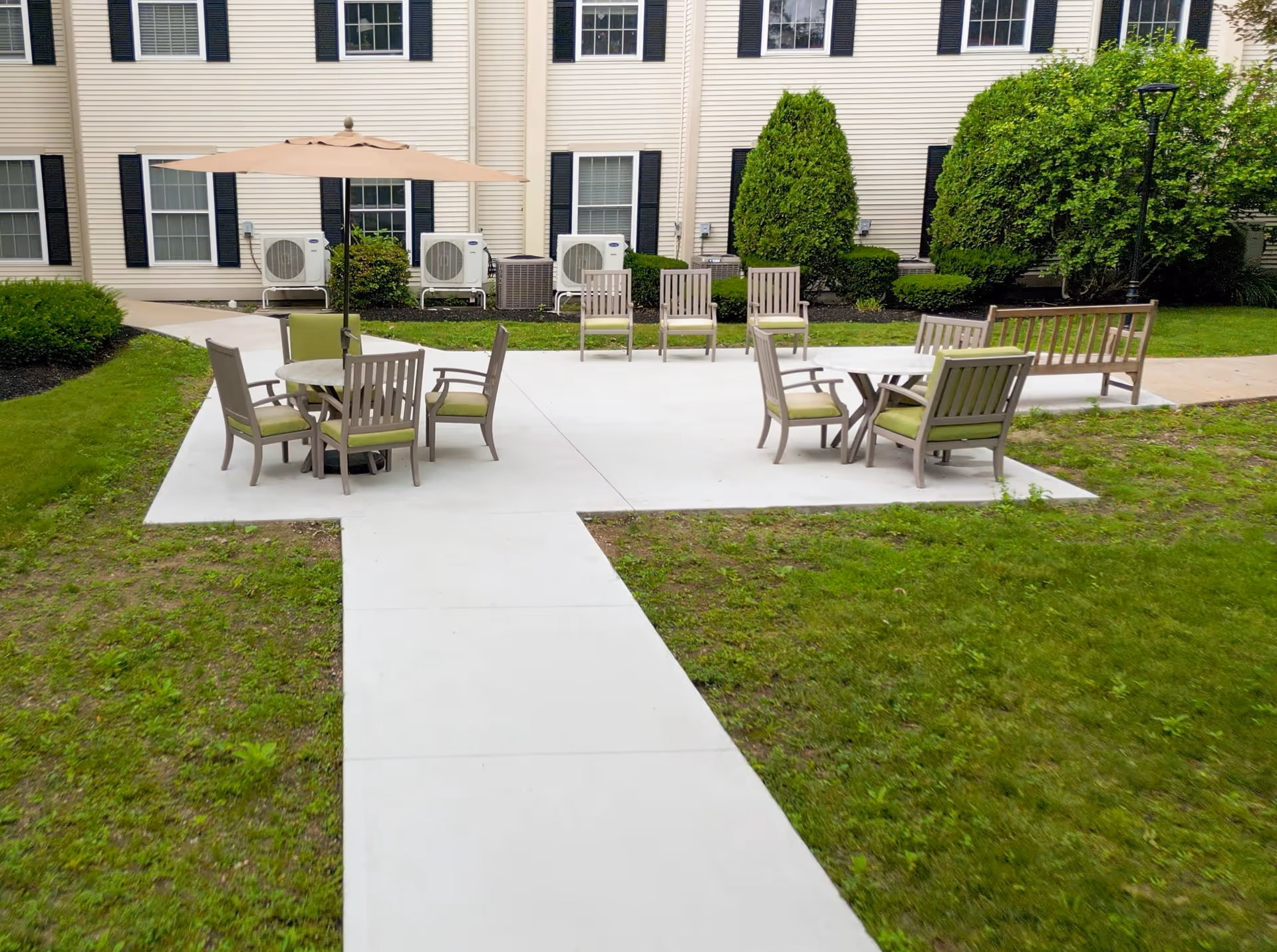 Outdoor patio area at Benchmark Senior Living at Waltham Crossings with multiple seating arrangements including chairs with green cushions, a round table with an umbrella, and benches. The patio is surrounded by green grass and bushes, with a beige building featuring windows and air conditioning units in the background.