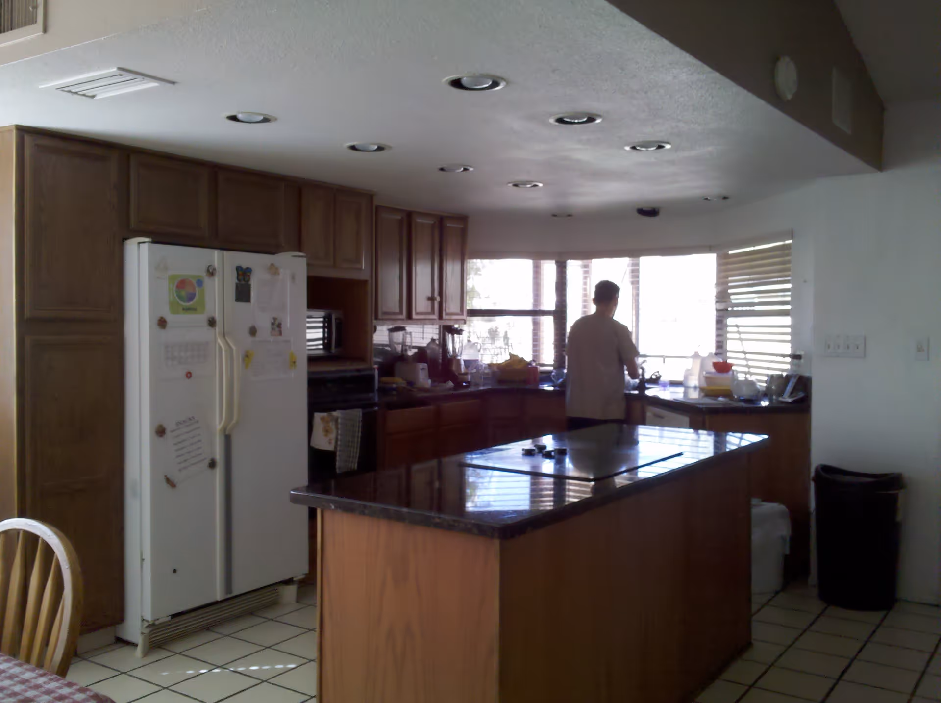 Kitchen interior with a central island, wood cabinets, a refrigerator, and a person standing at the sink by a bay window.
