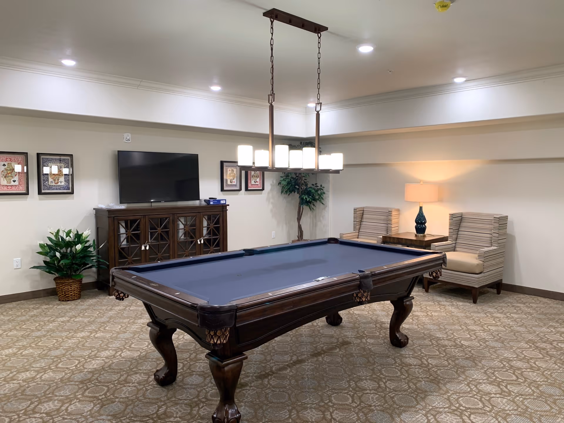 A well-lit game room featuring a dark wood pool table with a blue felt surface in the center. Behind the pool table is a wooden cabinet with glass doors, topped with a flat-screen TV. The walls are decorated with framed artwork. To the right, there are two striped armchairs with a small wooden side table and a lamp between them. A potted plant is placed near the cabinet, and the room has a patterned carpet and recessed ceiling lights.
