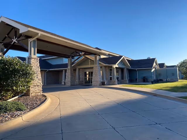 Front entrance of a single-story senior living facility with a covered porte-cochere, driveway, and clear blue sky.