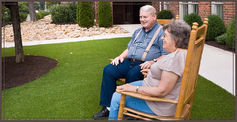 An elderly man and woman sitting and smiling on wooden rocking chairs on a green lawn outside a brick building with landscaping including rocks, bushes, and trees.