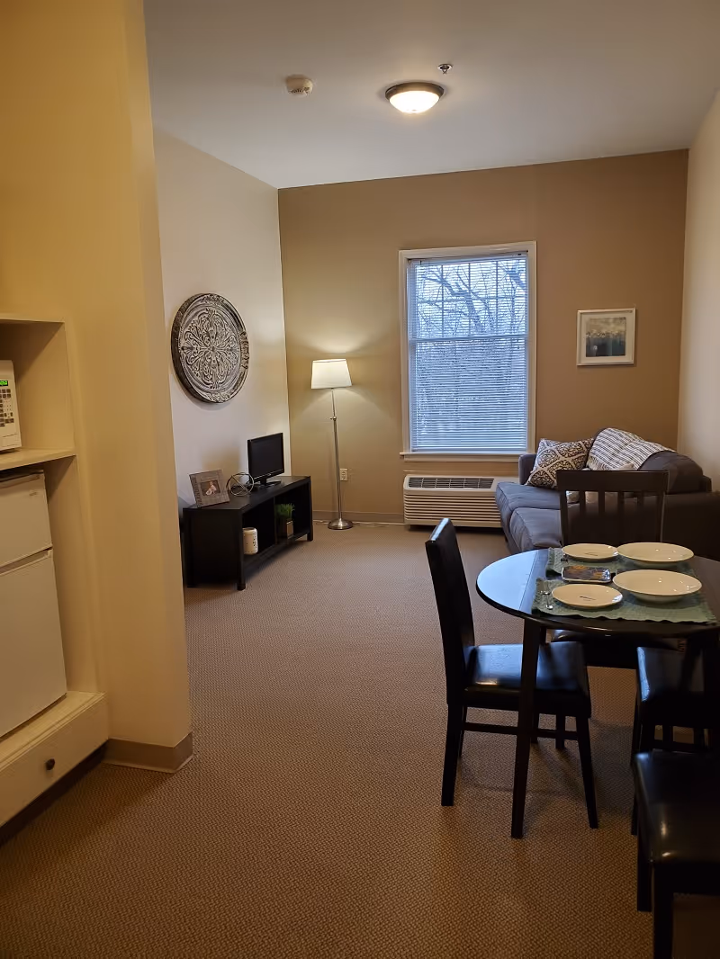 A cozy living room area with a gray couch adorned with patterned pillows, a floor lamp, a small TV on a black stand, and a window with blinds showing bare tree branches outside. In the foreground, there is a glass dining table set with four plates and four black chairs. To the left, a small kitchen area with a microwave and mini refrigerator is partially visible.