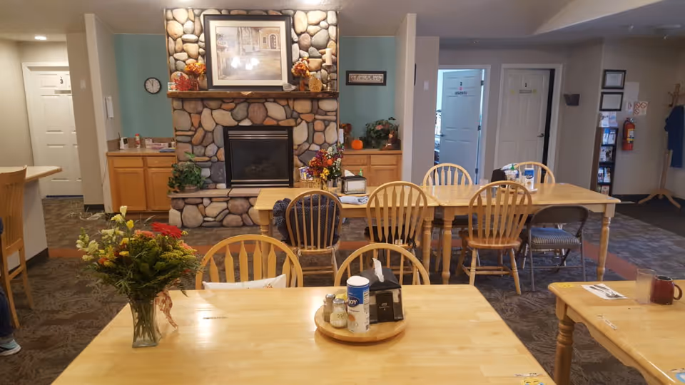 A cozy dining area in an assisted living facility with wooden tables and chairs arranged neatly. A stone fireplace with a framed picture and autumn decorations is centered on the back wall. There are flowers in a vase on the table in the foreground, and cabinets with plants and seasonal decor on either side of the fireplace. Doors and a clock are visible in the background.