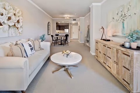 A bright and cozy living room area with a beige sofa adorned with decorative pillows, a round white coffee table, and a wooden sideboard with plants and a lamp. In the background, there is a dining table with four chairs and a kitchen area with dark cabinetry.