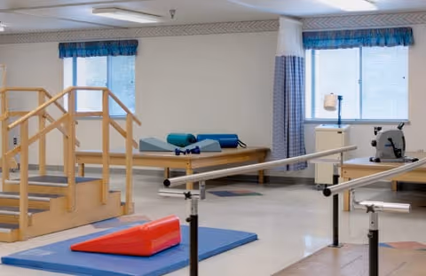 Physical therapy room with parallel bars for walking exercises, a wooden staircase for step training, padded mats, and exercise equipment near windows with blue valances and white curtains.