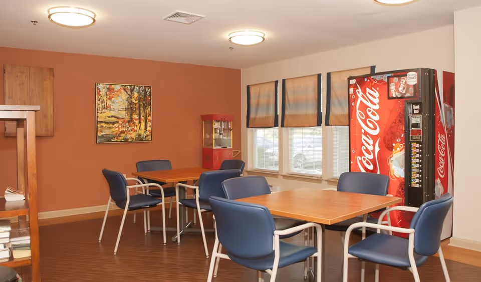 A small dining area with two wooden tables and blue cushioned chairs. There is a Coca-Cola vending machine and a popcorn machine against the wall. The walls are painted in a warm orange tone with a framed painting of a forest scene. Three windows with blinds allow natural light into the room.