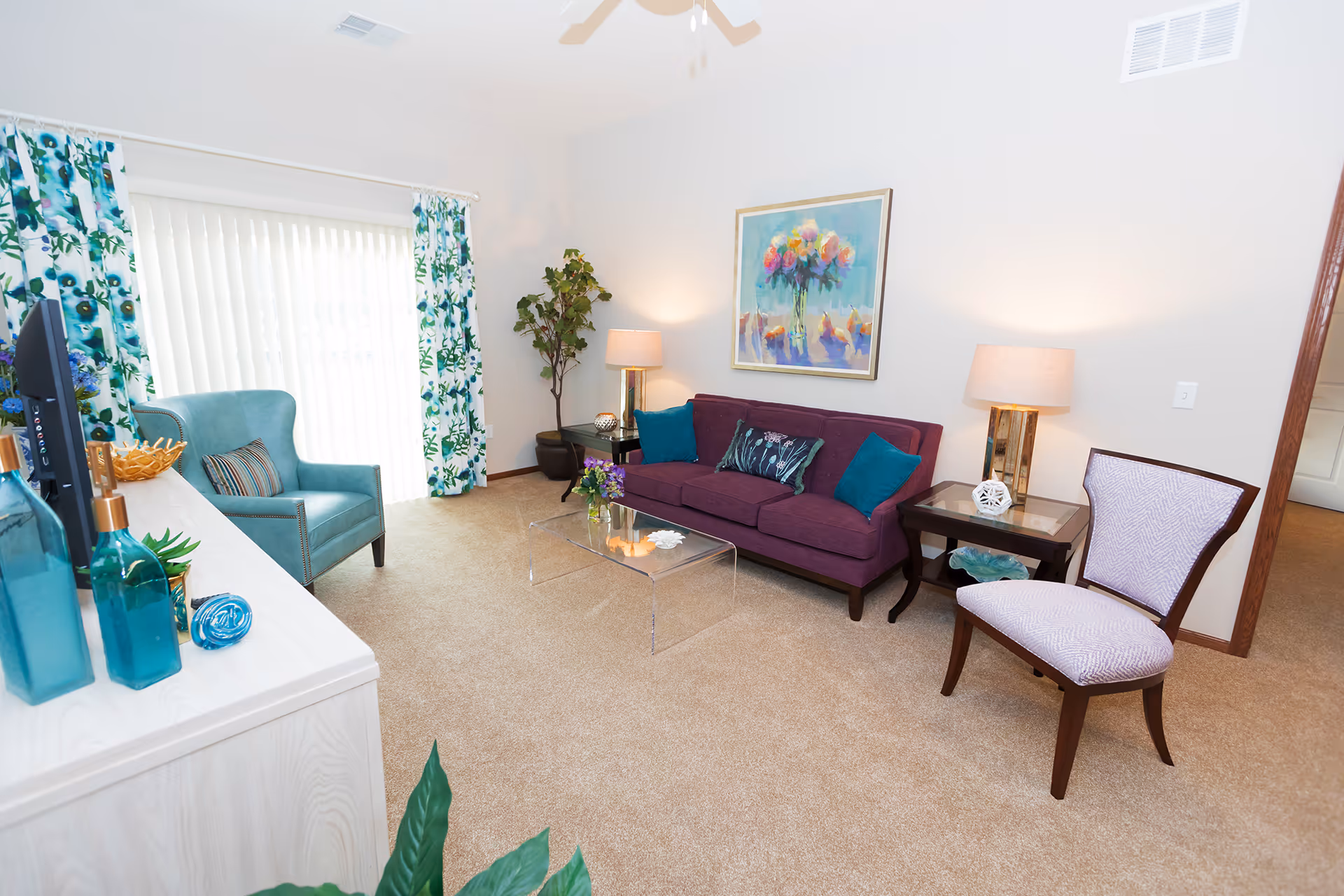 Bright, well-decorated living room featuring a purple sofa, teal armchair, accent chairs, a clear glass coffee table, and floral curtains.