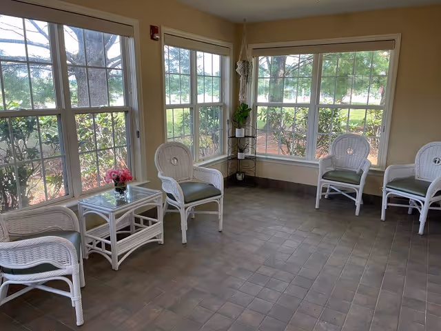 A bright room with large windows on two walls, featuring four white wicker chairs with green cushions arranged around a small glass-top wicker table with a vase of pink flowers. A small black metal plant stand with a green plant is in the corner.