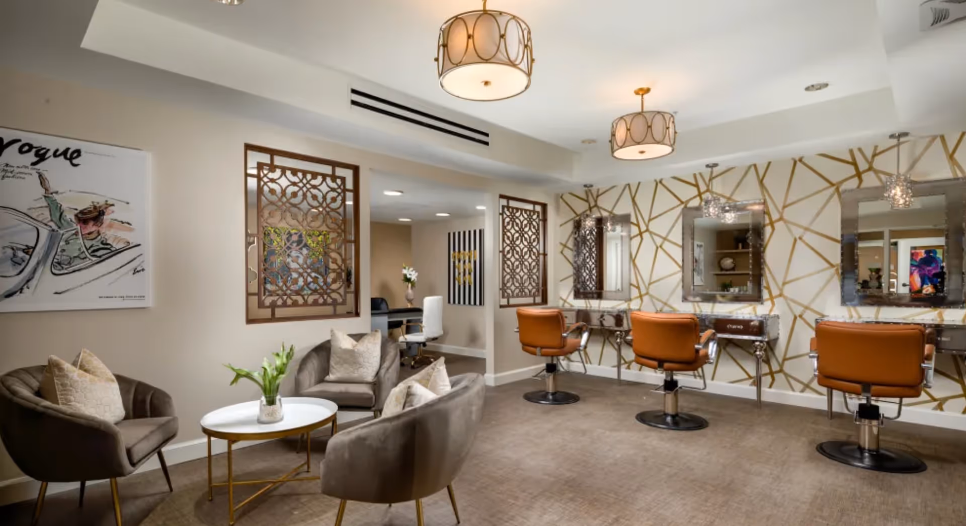 A stylish salon area in a senior living facility featuring three brown salon chairs in front of mirrors with decorative lighting. To the left, there is a cozy seating area with three gray armchairs around a small white and gold coffee table with a vase of flowers. The walls are decorated with gold geometric patterns and framed art, and the space is well-lit with ceiling lights.