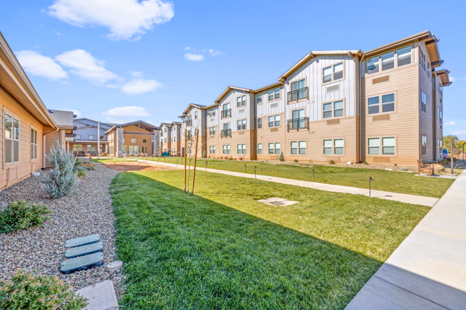 Grass courtyard and walkway between multi-story senior living buildings under a blue sky.