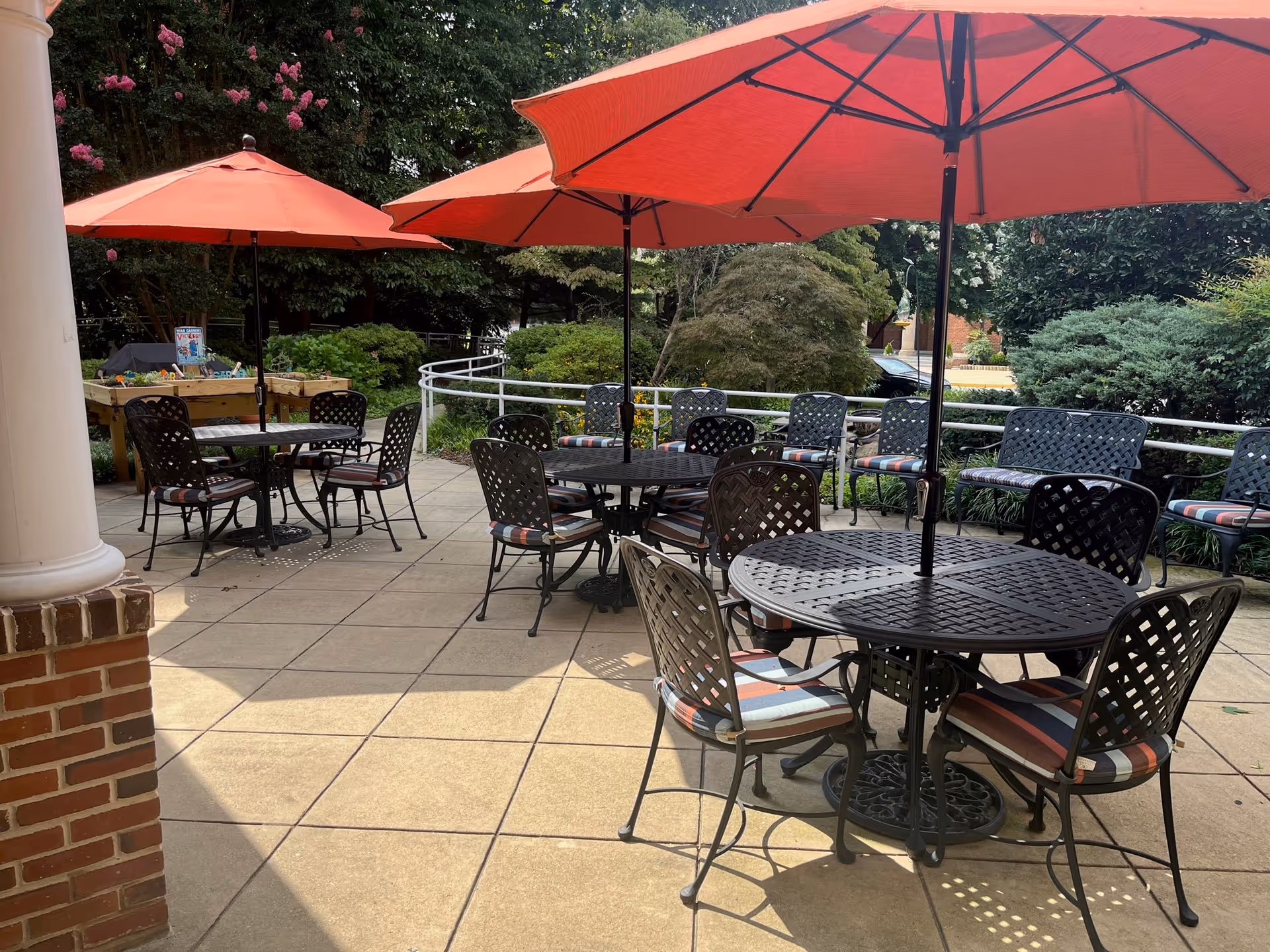 Outdoor patio with round metal tables and chairs shaded by red umbrellas overlooking landscaped greenery.